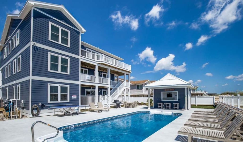 A multi-story blue house with a spacious backyard. A swimming pool, gazebo, and lounge chairs surround the pool. The sky is clear with white clouds.