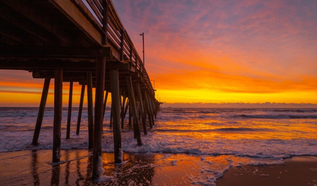 A serene sunset over a pier, with waves crashing against its structure, creating a picturesque coastal scene in Virginia Beach