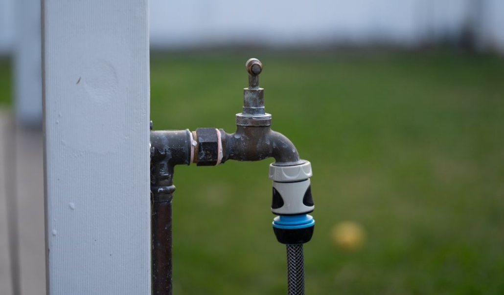 A close-up view of a water faucet mounted on a wooden fence in a backyard setting.