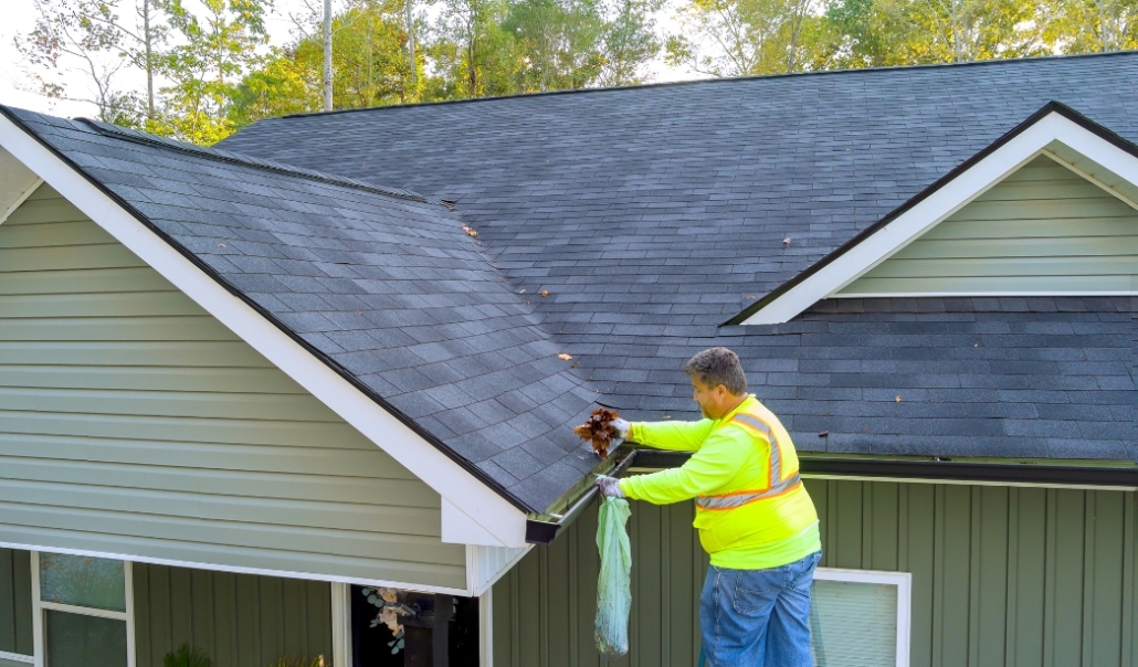 A man in a yellow vest is cleaning a house roof, focusing on seasonal maintenance of gutters and downspouts.
