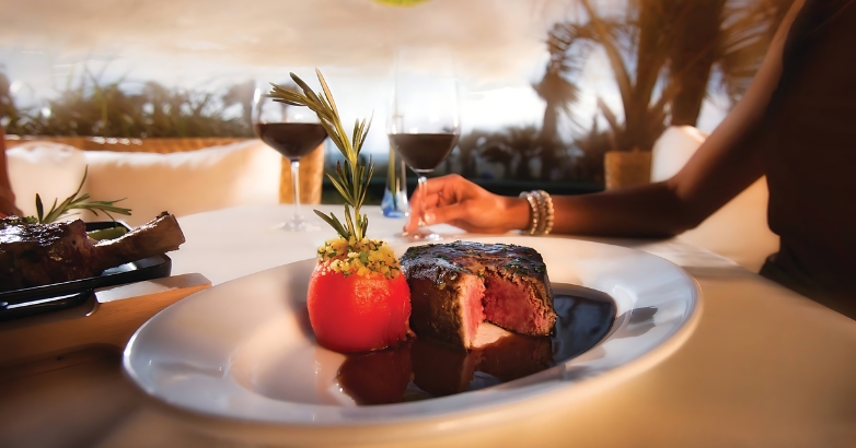 A woman presents a beautifully cooked steak on a plate, showcasing the vibrant dining scene in Salacia Prime Seafood and Steaks.