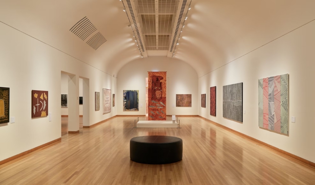 A view of an art gallery displaying paintings on the walls, accompanied by a black stool, reflecting Virginia Beach's artistic scene.