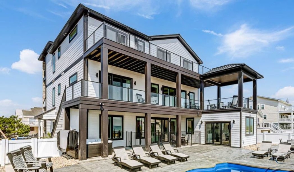 Modern three-story beach house with large glass balconies, black trim, and outdoor seating areas under a blue sky.