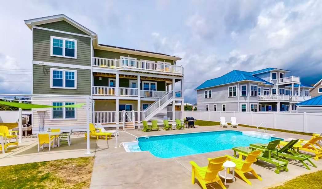 The image features a pool in front of a house with a deck, representing a Sandfiddler home in Pungo, Virginia Beach.